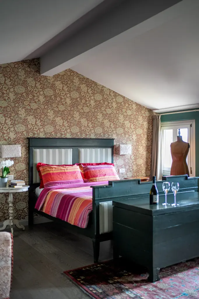 Letto matrimoniale con testata e cassapanca verde scuro, biancheria a righe rosa/arancio, carta da parati e manichino vicino alla finestra