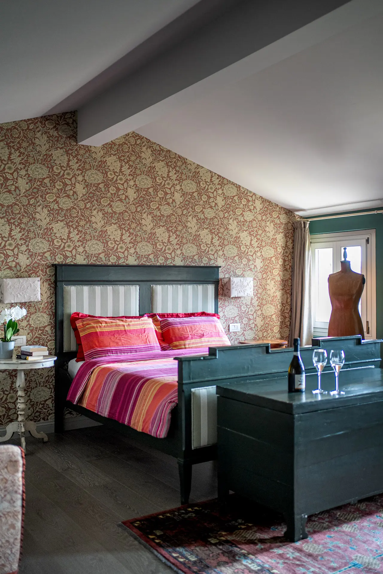Camera matrimoniale con testata e cassapanca verde scuro, biancheria a righe rosa/arancio, carta da parati e manichino vicino alla finestra