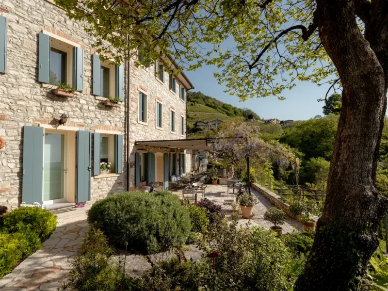 Facciata in pietra con persiane azzurro polvere, terrazza ombreggiata, piante e albero in primo piano