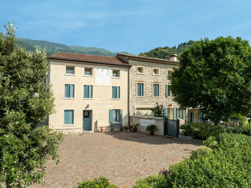 Facciata in pietra con cortile lastricato, persiane azzurro polvere, albero ombroso e colline sullo sfondo
